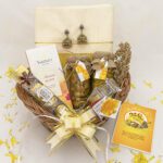 A beautifully arranged vishu gift basket for womens containing a bottle of banana chips, 4 Ferrero Rocher chocolates, Jimikki earrings, a greeting card, a Kerala saree, a personalized photo card, saffron and Sharkkaravaratty, and a bottle of Yardley Mist Perfume.