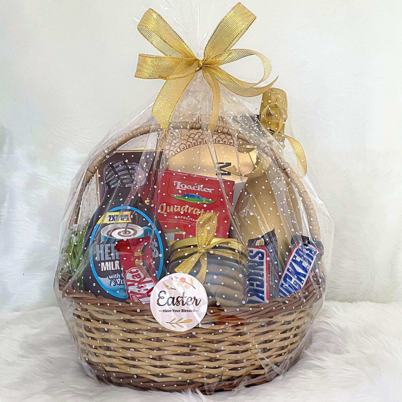 The featured Easter gift basket consists of Giacobazzi Non-Alcoholic Red Wine, Ferrero Rocher Moments Truffles, Kinder Bueno White Chocolate Bar With Coconut, Loacker Quadratini Napolitaner Waffles, Hershey's Milk Booster, chocolate cookies, KitKat, Snickers, and Gone Mad Choco Sticks.